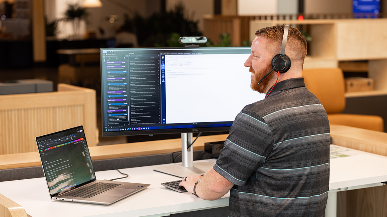 Man looking at a laptop with a larger monitor next to him, while wearing a headset and typing. 