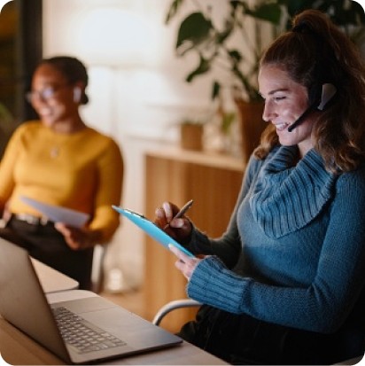 A smiling woman wearing a headset performing some action on a tablet, with a colleague in the background.