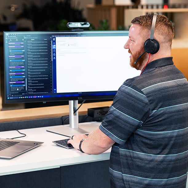 An associate stands at a desk, working on a computer with a large monitor and wearing headphones.