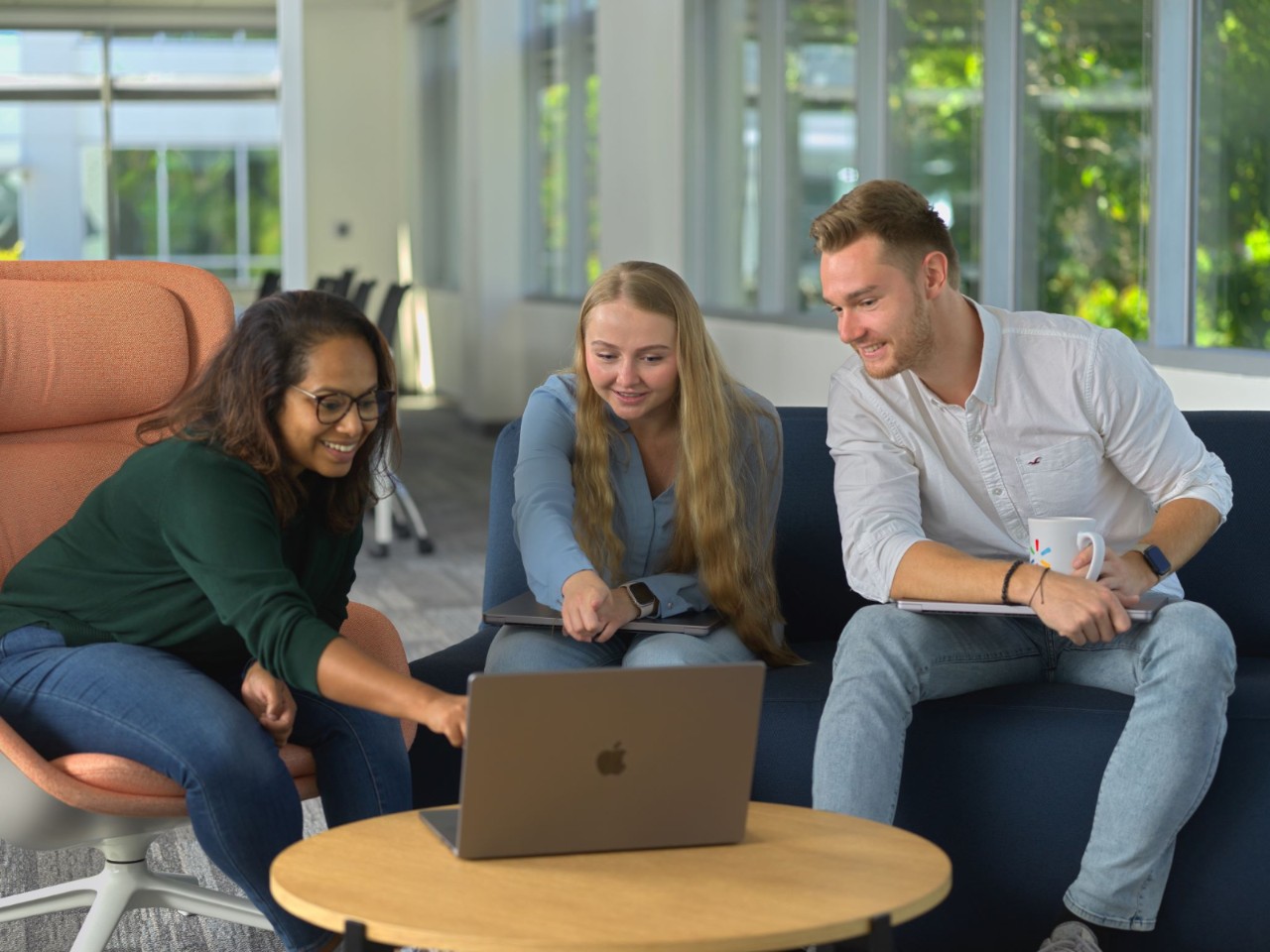 Three people sit on a couch and chair, looking at a laptop on a round wooden table in an office setting with large windows in the background.