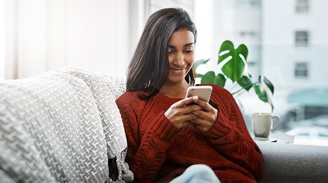 Image of a woman sitting inside a room on a couch. She is smiling and looking at the screen of a smartphone.